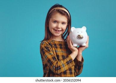 Cute Adorable Girl Kid Holding Money Box In Shape Of Pig Near Ear Looking Happy On Blue Studio Background.