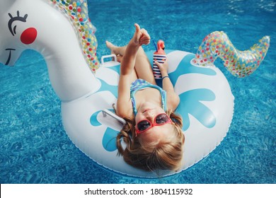 Cute Adorable Girl In Sunglasses With Drink Lying On Inflatable Ring Unicorn. Kid Child Enjoying Having Fun In Swimming Pool. Summer Outdoor Water Activity For Kids. View From Above Overhead.