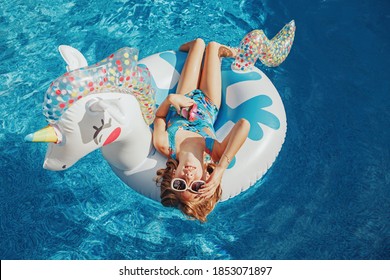 Cute Adorable Girl In Sunglasses With Drink Lying On Inflatable Ring Unicorn. Kid Child Enjoying Having Fun In Swimming Pool. Summer Outdoor Water Activity For Kids. View From Above Overhead.