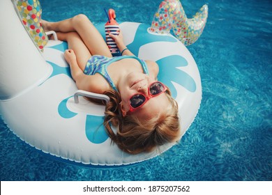 Cute Adorable Girl In Sunglasses With Drink Lying On Inflatable Ring Unicorn. Kid Child Enjoying Having Fun In Swimming Pool. Summer Outdoor Water Activity For Kids. View From Above Overhead.