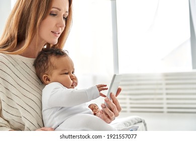 Cute adorable infant baby daughter learning using modern cell phone technology with mom working at home. Young caucasian mother holding smartphone and little mixed race african child daughter.