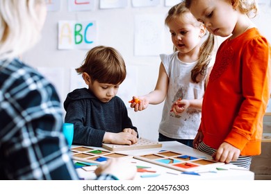 Cute, Adorable Little Boy And Two Girls Help To Do Logic Exercise With Figures And Shapes In Class Group. Teacher With Children In Kid Development Childcare Center For Preschoolers. Early Education