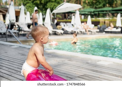 Cute adorable little caucasian blond toddler boy kid in diaper, inflatable ring sitting near poolside on wood flooring at pool edge before swimming. Happy child enjoy summer vacation travel family