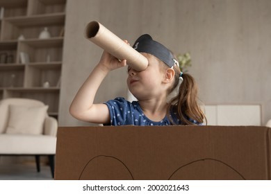 Cute adorable little girl in black pirate hat looking in spyglass, sitting in toy cardboard ship, preschool child kid wearing homemade costume holding cardboard tube as telescope, playing funny game
