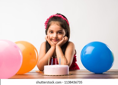 Cute adorable little Indian/asian small girl celebrating birthday while holding strawberry cake and blowing candles at table or standing isolated over white or red background 
