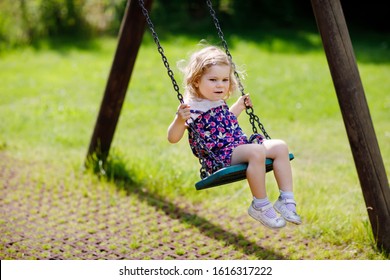 Cute adorable toddler girl swinging on outdoor playground. Happy smiling baby child sitting in chain swing. Active baby on sunny summer day outside