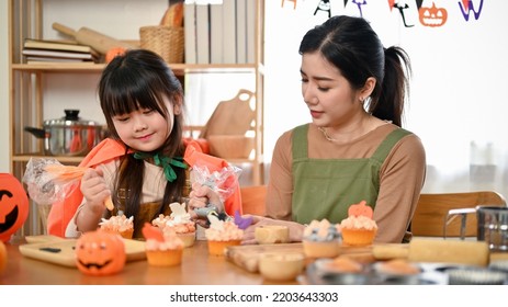 Cute And Adorable Young Asian Girl In Halloween Costume Enjoys Decorating And Making A Cupcake For Her Halloween Party At Home With Her Mom.