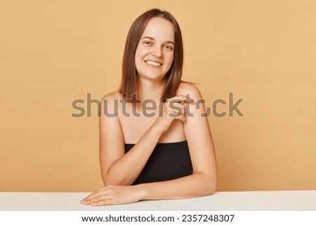 Cute adorable young happy woman wearing black top looking at camera posing isolated over beige background expressing happiness smiling with joyful face.