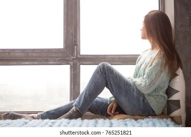 Cute adult young woman sitting near window in casual clothes (sweater and jeans), looking sideways thoughtful. Copy space - Shutterstock ID 765251821