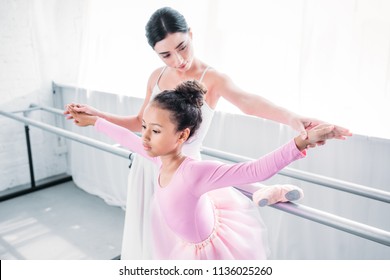 cute african american child in pink tutu exercising with teacher in ballet school