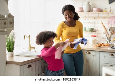 Cute African American Girl Helping Mother With Household, Wiping Dishes In Kitchen After Washing. Adorable Black Little Helper Child Enjoying Spending Time With Mom At Home, Learning Housekeeping
