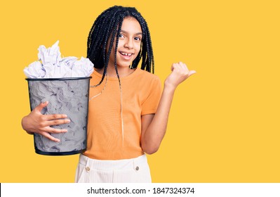 Cute African American Girl Holding Paper Bin Full Of Crumpled Papers Pointing Thumb Up To The Side Smiling Happy With Open Mouth 