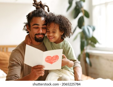 Cute African American Little Boy Son Giving His Father Handmade Greeting Postcard With Drawn Red Heart And Congratulating Dad With Fathers Day At Home. Family Celebration Concept