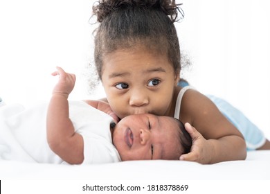 Cute African American little girl kissing on newborn baby cheek on white bed at home. Little girl takes care of infant baby with kindly