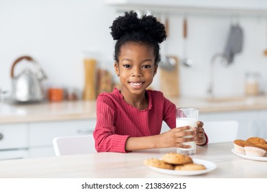 Cute african american little girl drinking milk, smiling at camera. Adorable black child sitting at table, having snack, holding glass of milk, eating cookies, kitchen interior, copy space
