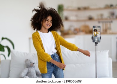 Cute African American Preteen Girl With Bushy Hair In Casual Outfit Recording Video While Dancing, Showing Dance Moves, Using Cell Phone On Tripod, Home Interior, Copy Space. Kids Lifestyle Concept