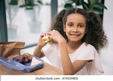 Cute African American Schoolgirl Eating Sandwich And Smiling At Camera At Lunch In School