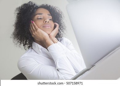 Cute African American woman holding her head with both hands and looking at her laptop screen in office.