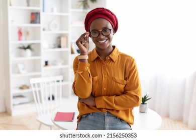 Cute African American Young Lady In Casual Outfit And Red Turban Posing At Workplace, Standing By Desk, Touching Her Eyeglasses And Smiling At Camera, Copy Space. Career For Millennials