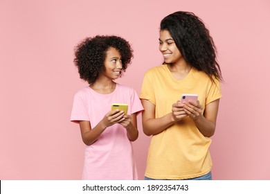 Cute African American Young Woman Little Kid Girl Sisters In T-shirts Using Mobile Cell Phone Typing Sms Message Looking At Each Other Isolated On Pink Background Studio Portrait. Family Day Concept