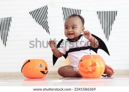 Cute African baby kid dressing up vampire fancy Halloween costume with black bat wings, cheerful little cute child holds Halloween pumpkin to play trick or treat at party. Happy Halloween celebration.