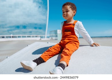 Cute african baby toddler in a orange overalls sits on a stone bench by the sea at city. Happy girl playing in summer alone. - Powered by Shutterstock