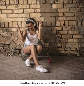 Cute African Black Girl Sit Near Cracked Grunge Yellow Brick Wall With Big White Headphones Female Hold In Head Mobile Phone Portrait Of Young Adult Attractive Afro American Woman Listening Music