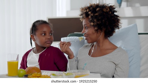 Cute African Girl Visiting Sick Mother In Hospital At Lunch Time. Portrait Of Afro Woman Patient With Nasal Tube Eating Meal In Hospital Bed And Talking To Little Daughter Visitor