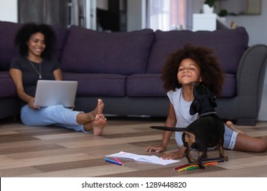 Cute African Kid Girl Playing With Dog Spending Time With Mom In Living Room, Black Mother And Child Daughter Having Fun With Dachshund On Floor, Happy Mom And Child Enjoy Leisure At Home With Pet