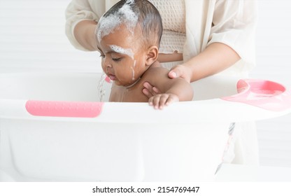 Cute African Newborn Baby Bathing In Bathtub With Soap Bubbles On Head And Body. Asian Young Mother Washing Her Little Daughter In Warm Water. Newborn Baby Cleanliness Care Concept