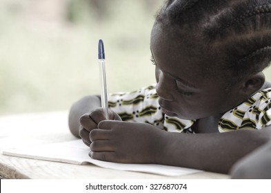 Cute African Schoolgirl Doing Her Homework At School: Beautiful Black Girl Writing And Learning Activity With A Blue Pen. She's Sitting In Her Desk Doing Her Calculating Work In Her Exercise Book.