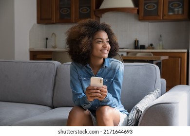 Cute African teenage girl resting on cozy sofa holds smartphone staring aside looks dreamy, distracted from modern device usage, consider purchase feels satisfied spend free time at home using tech - Shutterstock ID 2171644693