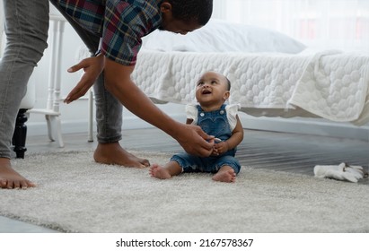 Cute African Toddler Baby Smiling To Father Who Standing And Bow Down Give Her Sweets While Sitting On Carpet On Floor At Bedroom. Relationship Of Dad And Little Daughter Concept