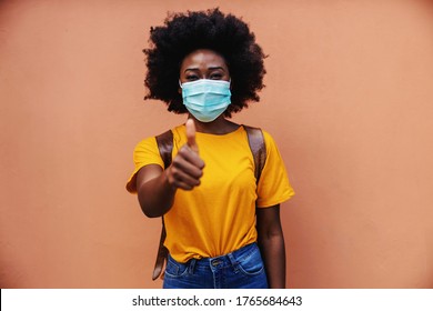 Cute African Woman Standing Outdoors With Mask On Her Face And Showing Thumbs Up. Covid-19 Outbreak.