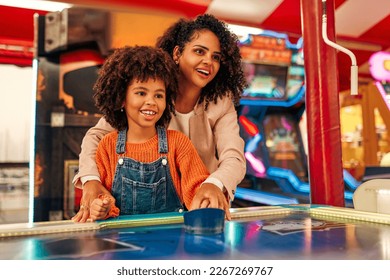 A cute African-American child with afro curls with her mother playing air hockey at an amusement park and carousel on her day off in the evening.