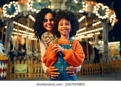 A cute African-American girl with an Afro hairstyle with her mother eating a colorful lollipop standing against the background of a carousel with horses in the evening at an amusement park or circus.