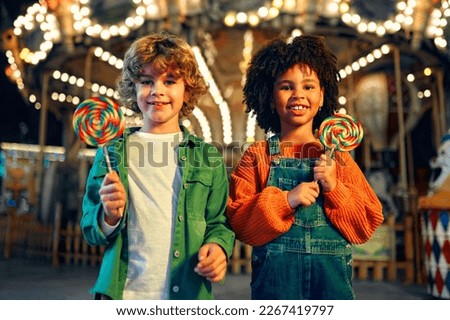 A cute African-American girl with an Afro hairstyle and caucasian boy eating a colorful lollipop standing against the background of a carousel with horses in the evening at an amusement park or circus