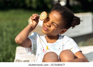 A Cute African-American Girl Covers One Eye With A Bright Fruit Lollipop In The Park On A Summer Day.Summer Holidays,diverse People Concept.