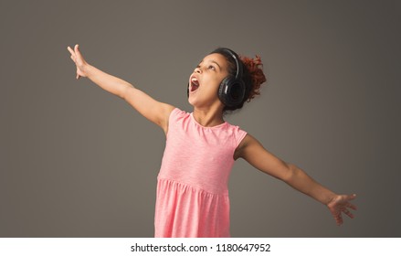 Cute african-american girl singing in headphones at gray studio background