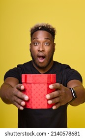 A Cute African-American Man Opens His Mouth And Hands The Birthday Boy A Gift In A Red Paper Box. A Curly Black Man Is Worried, Surprises His Girlfriend By Opening His Mouth Wide And Opening His Eyes