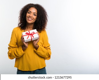 Cute african-american young woman in yellow autumn top holding gift box with red ribbon and bow on white wall background. Girl smiling, she is happy to get present