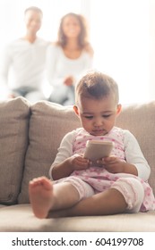 Cute Afro American Baby Is Using A Smart Phone While Sitting On The Couch, Her Parent In The Background