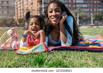 Cute Afro Girl Lying Down In The Grass In Colorful Picnic Blanket With Her Mom And Looking At Camera.