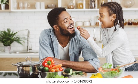 Cute afro kid girl feeding her father while they cooking fresh salad. Soul kitchen concept