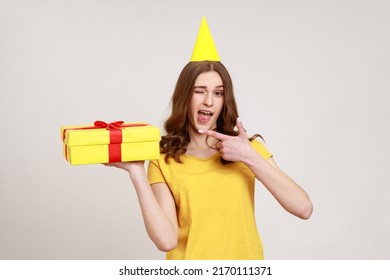 Cute amazed teenage girl with party cone in yellow T-shirt pointing at gift box and winking to camera, showing awesome birthday present. Indoor studio shot isolated on gray background.