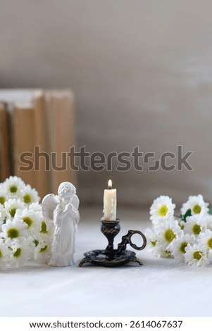 cute angel figurine, burning candle, white flowers and books on table. symbol of Christianity religion, faith in God, prayer, memory. christian home altar. soft focus
