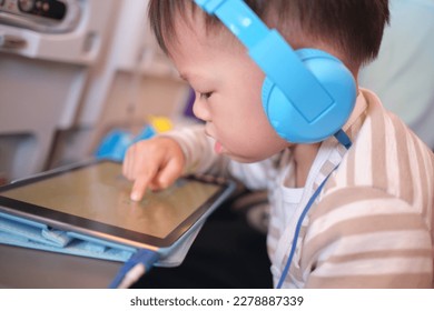 Cute Asian 3 years old toddler boy child wearing headphones using tablet pc watching cartoons, playing game during flight on airplane. Happy Flying with children concept, Soft and selective focus - Powered by Shutterstock