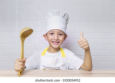 Cute Asian Boy With Apron And Chef Hat Enjoy Preparing Food In Cooking Class On White Brick Background Using For Kid And Family Concept, Cooking Class Or Food And Beverage Content. 