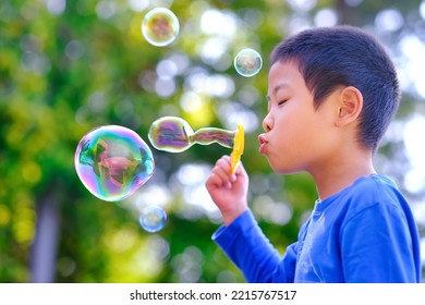 Cute Asian boy closing eye and blowing bubbles at outdoor against blurred green tree background. Happy childhood fun play time. - Powered by Shutterstock