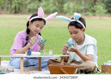 Cute Asian boy and girl with bunny ears painting egg with paintbrush while sitting together on green grass meadow in garden. Kids celebrating Easter holiday outdoor. Happy children in park on Easter - Powered by Shutterstock